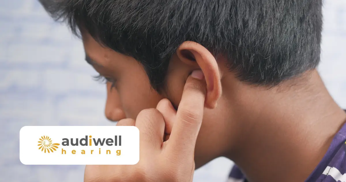 Young person pressing a finger behind their ear, a common sign of tinnitus, with the Audiwell Hearing logo